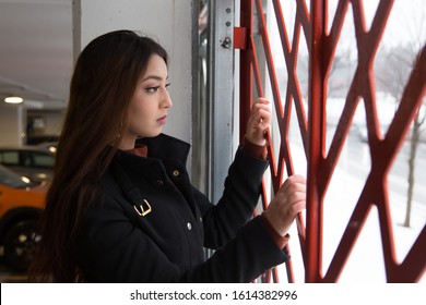 Medium Horizontal View Of Pretty Brunette Woman With Waist Length Hair Wearing Black Coat Looking Out Of The Window Of An Indoor Parking Building 