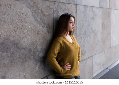 Medium Horizontal View Of Pretty Brunette Woman With Waist Length Hair In Mustard Coloured Sweater Leaning On Stone Wall With Hands On Hips While Looking Ahead With Stern Expression