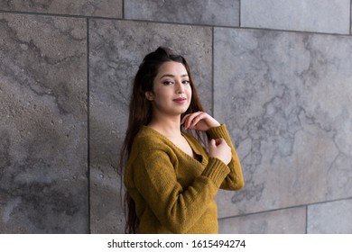 Medium Horizontal View Of Pretty Smiling Brunette Woman With Waist Length Hair Wearing A Mustard Coloured Sweater And Standing In Front Of Stone Wall