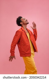 Medium image of young American guy pretending to move like robot against pink background. Boy with trendy afro haircut in cool outfit of lemon vintage jeans with tank top and orange jacket.