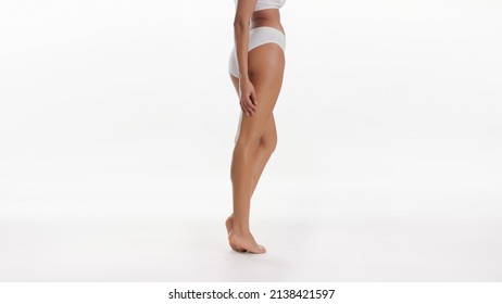 Medium Long Shot Of African American Good-looking Slim Woman In White Underwear Touches Her Thigh Standing On Tiptoes On White Background | Smooth Legs Skin Concept