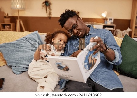 Medium long shot of African American dad wearing glasses reading fairy tales to sleepy little daughter while sitting on couch