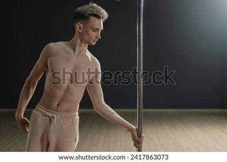 Medium long shot of bare-chested Caucasian man gripping pylon preparing for dance stunt in studio