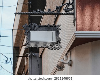 A medium, low-angle shot capturing a blank, ornamental European-style hanging storefront sign mounted on the exterior wall of a building. The rectangular sign features a blank white center panel suited for copy, framed by a highly detailed, textured silver-gray metallic border carved with decorative roses and leaf vines. It hangs by short chains from a wrought-iron scrollwork bracket attached to a beige-tiled and red-plastered building corner. A white outdoor spotlight or security camera base is mounted directly underneath the sign. In the blurred background, overhead utility power lines and adjacent urban buildings are visible against a clear blue sky under bright daytime conditions.
