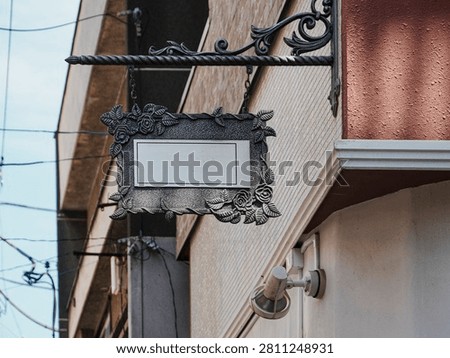 A medium, low-angle shot capturing a blank, ornamental European-style hanging storefront sign mounted on the exterior wall of a building. The rectangular sign features a blank white center panel suited for copy, framed by a highly detailed, textured silver-gray metallic border carved with decorative roses and leaf vines. It hangs by short chains from a wrought-iron scrollwork bracket attached to a beige-tiled and red-plastered building corner. A white outdoor spotlight or security camera base is mounted directly underneath the sign. In the blurred background, overhead utility power lines and adjacent urban buildings are visible against a clear blue sky under bright daytime conditions.