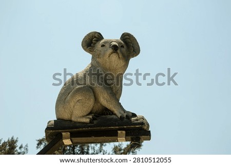 A medium low-angle shot of a realistic grey koala bear sculpture or statue perched on top of a weathered wooden post. The decorative animal figurine features detailed fur textures, large rounded ears, and a prominent black nose, looking upwards into the distance. Set against a clear blue sky background with tree tips visible in the corner, this image is perfect for themes of zoo signage, Australian landmarks, park decorations, and tourism concepts.