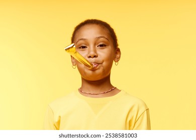 Medium portrait shot of happy Black girl blowing in party horn wearing yellow t shirt while standing against background of matching color in studio, copy space