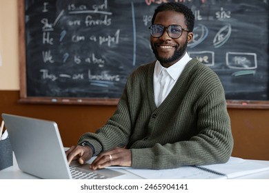 Medium portrait of young African American male teacher of English wearing eyeglasses sitting at desk in classroom smiling at camera while using laptop - Powered by Shutterstock