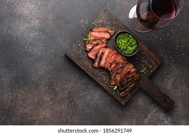 Medium Rare Steak Sliced On Black Cutting Board With Pesto Sauce And Glass Of Red Wine. Dark Brown Food Background