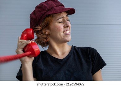 Medium Short Shot Of Latin American Young Woman (22) With Cap And Red Hair Uncomfortable Because Of The Screams She Hears From The Conversation Over The Red Retro Handset. Vintage Technology Concept.