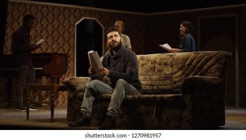 Medium shot of an actor reciting his lines on a sofa while other actors rehearsing in the background