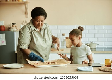 Medium shot of African American grandmother holding baking tray with tasty biscuits cooking homemade pastry together with curious little granddaughter at beige cozy kitchen - Powered by Shutterstock