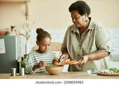 Medium shot of African American little girl carefully mixing green lettuce leaves in wooden bowl, while helping grandmother with cooking salad with fresh tomatoes at beige kitchen - Powered by Shutterstock