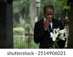 Medium shot of African American man in funeral black suit grieving at graveyard wiping tears and holding white lilies on sorrowful rainy day, copy space