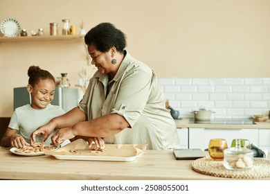 Medium shot of African American smiling girl baking delicious homemade cookies together with grandma while putting crispy biscuits from baking tray on plate at cozy beige kitchen, copy space - Powered by Shutterstock