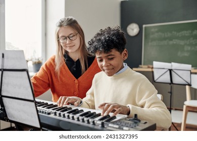 Medium shot of African American teen boy playing electronic piano at music class, his young Caucasian teacher sitting next to him - Powered by Shutterstock