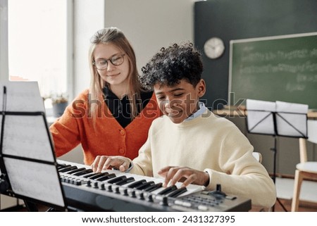 Medium shot of African American teen boy playing electronic piano at music class, his young Caucasian teacher sitting next to him