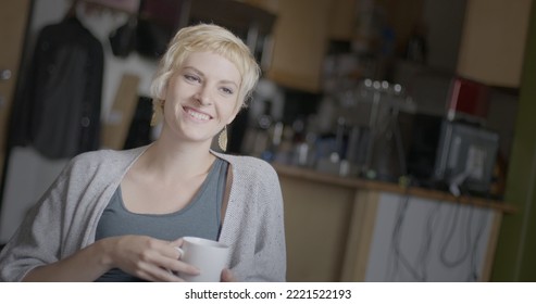 Medium Shot Of Attractive Young Woman With Short Blonde Hair, Smiling And Drinking Coffee From A White Mug In An Urban Loft