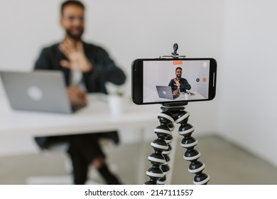 Medium Shot Of Black Male Through Focused Camera Phone On Tripod And Sitting At Desk With Laptop And Explaining Something On Camera While Filming Vlog In Cozy Studio Office