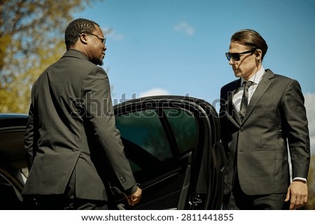 Medium shot of Black middle aged man entering car while Caucasian young adult chauffeur assisting beside open door, conveying executive transport service. Ideal for business travel marketing