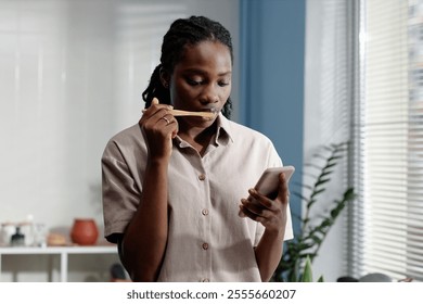 Medium shot of Black woman glued to phone screen while brushing teeth during self care routine in bathroom on Sunday morning, copy space - Powered by Shutterstock