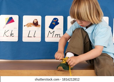 Medium shot of a boy tying his shoe lace while sitting on a wooden storage cabinet in his classroom. Stock Photo