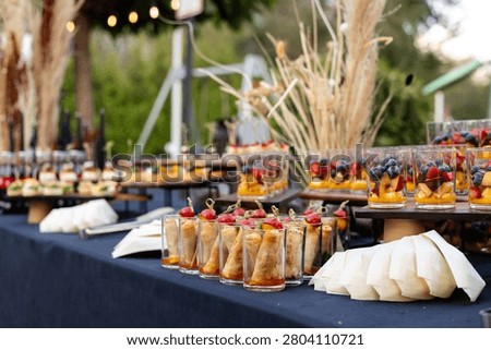 Medium shot of a buffet table with transparent glasses of pancake rolls with cherry tomatoes and fruit salads with berries, decorated with pampas grass