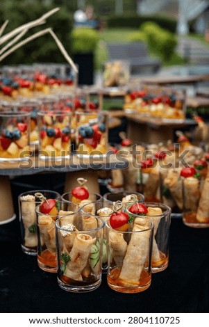 Medium shot of a buffet table with transparent glasses containing pancake rolls with cherry tomatoes on skewers and fruit salad 