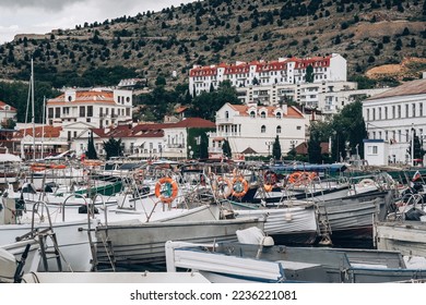 A medium shot captures a densely packed marina filled with various small boats, many with orange life preservers. White buildings with red roofs line the hillside in the background under a cloudy sky.