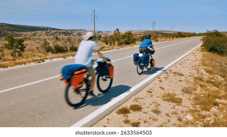 A medium shot captures two cyclists riding down a long, empty road. The person in the foreground, wearing a white hat and blue and white shirt, rides a bike with orange and blue bags.
