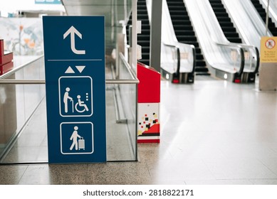 A medium shot of a clear blue elevator directional sign within a train station or public building. The sign features universal pictograms for a person in a wheelchair, an elderly person with a walking stick, and a person with luggage, along with up and down arrows. In the background, two operating escalators are visible next to a polished floor. The scene is orderly and modern.