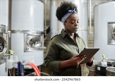 Medium shot of concentrated female African American supervisor using tablet computer inspecting production process in workshop of cider or beer factory, copy space - Powered by Shutterstock