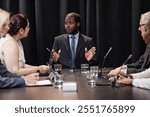 Medium shot of confident African American male presidential candidate speaking with political party committee developing campaign strategy while sitting at table against black curtains, copy space