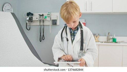 Medium Shot Of A Cute Little Boy Dressed Up As A Doctor In An Oversized Coat Using A Tablet Computer In A Medical Exam Room