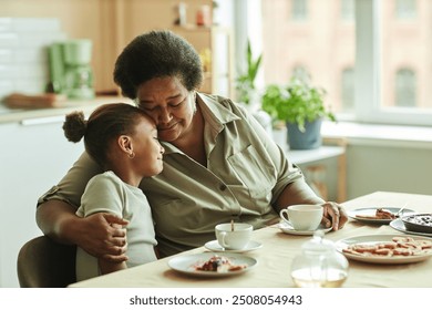 Medium shot of doting grandma gently embracing little granddaughter with love and affection while having tea sitting at table against window at sunlit kitchen, copy space - Powered by Shutterstock