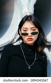 Medium shot of fashionista girl in black sweater and sunglasses standing in front of big concrete tube in sunlight.