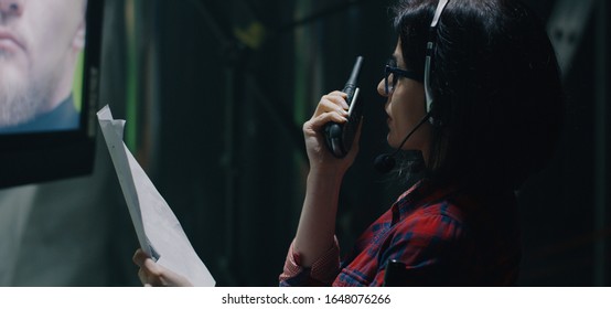 Medium shot of a female director watching an actor on screen and giving instructions on walkie-talkie