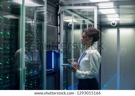 Medium shot of female technician working on a tablet in a data center full of rack servers running diagnostics and maintenance on the system