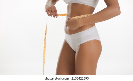 Medium shot of fit African American woman in underwear measures her waistline using tape on white background | Body health care concept