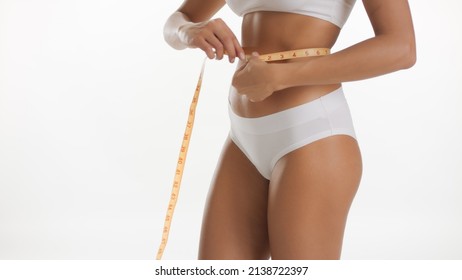 Medium shot of fit African American woman measures her waistline using measuring tape on white background | Body health care concept