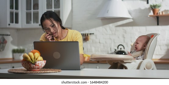 Medium Shot Of A Frustrated Young Mother Trying To Work At Home In The Kitchen With Crying Baby Next To Her