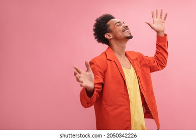 Medium shot of happy African young and smiling man with closed eyes with copy space. Well dressed in fall colors with arms raised at his sides against solid pink background.