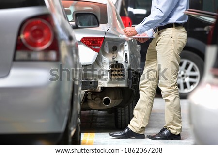 Medium shot of an insurance assessor making notes on a clipboard while inspecting a damaged vehicle at a garage.