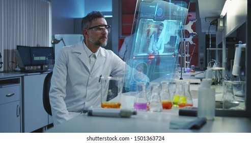 Medium shot of a male scientist using a holographic display screen for video call with other male medical specialist
