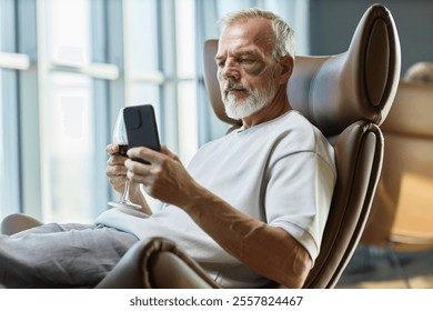 Medium shot of mature businessman taking care of face skin with eye patches, while text messaging on mobile phone over glass of red wine in comfy armchair at home - Powered by Shutterstock