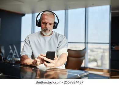 Medium shot of mature man with well groomed white beard listening to music in headphones while using mobile phone at table in contemporary hotel room, copy space - Powered by Shutterstock