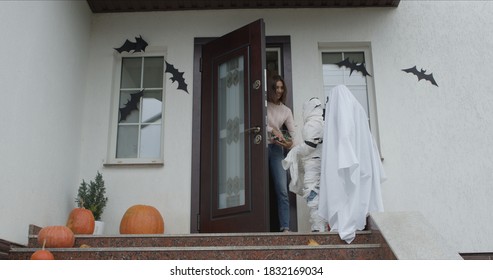 Medium Shot Of Mummy And Ghost Coming For Candy. Two Kids Wearing Cool Halloween Costumes Knocking On House Door. The Hostess Opens Doors And Shares Sweets.