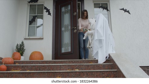 Medium Shot Of Mummy And Ghost Coming For Candy. Two Kids Wearing Cool Halloween Costumes Knocking On House Door. The Hostess Opens Doors And Shares Sweets.