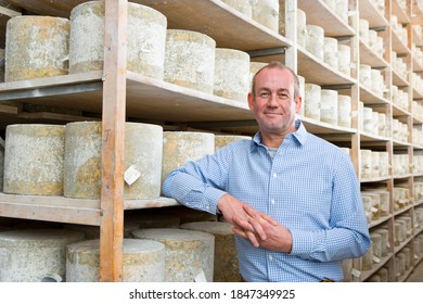 A medium shot of a owner standing in a cellar and smiling with aged cheddar cheese wheels in background. - Shutterstock ID 1847349925
