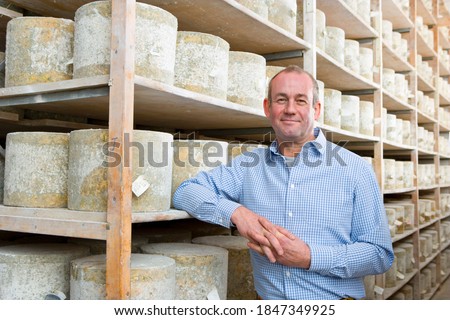 A medium shot of a owner standing in a cellar and smiling with aged cheddar cheese wheels in background.